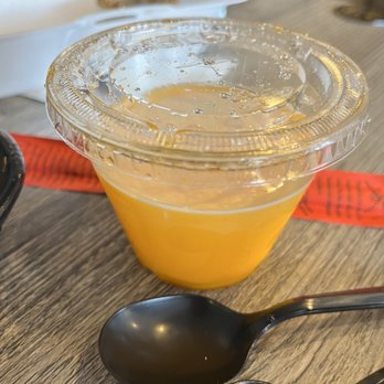 IT DIMSUM - Small Mango Pudding - A small, transparent plastic cup filled with mango pudding, placed on a wooden table with two black spoons nearby. - Dim Sum Near Me - Albuquerque, New Mexico