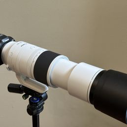 A Canon camera with a long, white telephoto lens attached is mounted on a tripod stand. The lens features a black focus ring and a large black lens hood at the front. The setup is placed indoors against a plain beige wall, indicating a photography or videography environment.