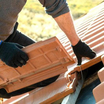 Ray Roofing - A person wearing black gloves is installing or repairing clay roof tiles on a sloped roof. The individual is holding a tile and appears to be adjusting or placing it. The roof has a series of curved tiles, and the background shows trees and sunlight, indicating an outdoor setting during daytime. - Roofing Near Me - Hackensack, New Jersey