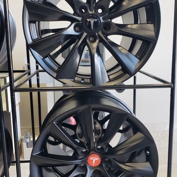 Tires 23 - Two black Tesla alloy wheels are displayed on a metal rack, with the top wheel showing a Tesla logo in the center and the bottom wheel featuring a red Tesla emblem. The wheels are positioned vertically, one above the other, in a retail or display setting. The background includes a tiled floor and some additional objects, indicating an indoor environment for automotive accessories. - Auto Parts & Supplies Near Me - Markham, Ontario