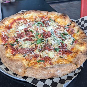 Hawt Pizza Co. - Yummy ham and pineapple pizza - A pizza with a thick, golden-brown crust, topped with bacon, chopped green herbs, cheese, and tomato sauce. The pizza is served on checkered paper, placed on a black table in a restaurant setting with glass windows in the background. A person's hand is visible in the top left corner, holding a napkin. - Pizza Near Me - Albuquerque, New Mexico