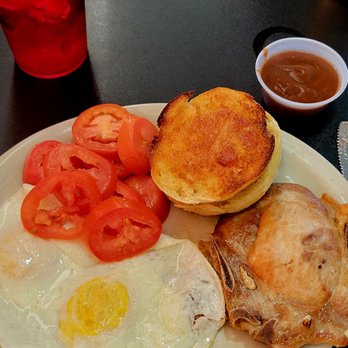 1431 Cafe - Porkchops with 2 fried eggs, tomatos, and an English muffin - A breakfast meal featuring fried eggs with sliced tomatoes, a biscuit, a piece of fried chicken, and a small cup of gravy on a plate. - American Near Me - Cedar Park, Texas