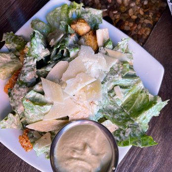 The Kosmos - Caesar Salad - A Caesar salad featuring chopped romaine lettuce, shaved parmesan cheese, croutons, and creamy Caesar dressing, served on a white plate. - Bars Near Me - Albuquerque, New Mexico