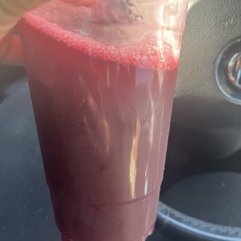 Jamaican Kitchen - Sorrell - A clear plastic cup contains a dark red liquid, likely juice or soda, with bubbles on the surface. The cup is held inside a vehicle, with part of the dashboard visible. - Caribbean Near Me - Vernon, Connecticut