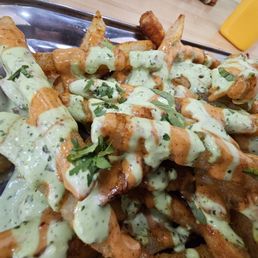 A metal tray contains a serving of French fries topped with green sauce, herbs, and drizzled with reddish sauce. The fries are garnished with chopped herbs, and the tray is placed on a wooden table with a yellow drink container visible in the background.