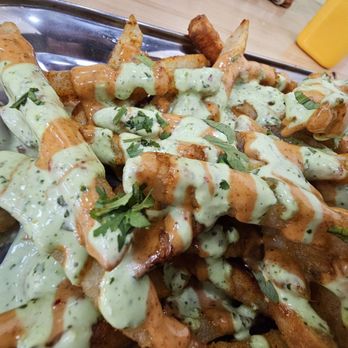 Moti Cafe - A metal tray contains a serving of French fries topped with green sauce, herbs, and drizzled with reddish sauce. The fries are garnished with chopped herbs, and the tray is placed on a wooden table with a yellow drink container visible in the background. - Indian Near Me - Glenview, Illinois