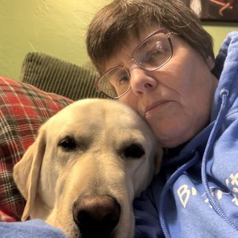 Curious K9 - Me and my dog Glacier - A woman with short brown hair and glasses is lying on a couch, resting her face against a yellow Labrador Retriever. She is wearing a blue hoodie, and the dog appears relaxed with its eyes partially closed. The background includes a green wall, a plaid pillow, and a framed picture hanging on the wall. The setting suggests a cozy indoor environment, likely a living room. - Pet Training Near Me