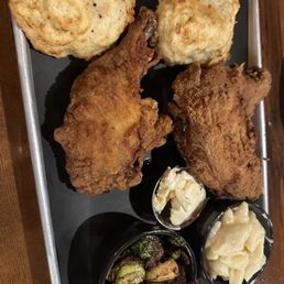 Fried Chicken - The image shows a metal tray containing fried chicken pieces, two biscuits, mashed potatoes with butter, and a side of cooked greens, likely collard or mustard greens. The fried chicken has a crispy, golden-brown coating, and the biscuits are flaky and slightly browned. The mashed potatoes are topped with a pat of butter, and the greens appear sautéed or simmered. The presentation suggests a Southern-style meal served in a casual dining setting.