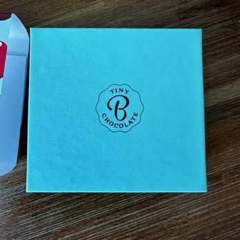 Tinyb Chocolate - A light blue, square-shaped box featuring the Tiny Chocolate logo in black, which includes a stylized 'B' and the words 'Tiny Chocolate' around it. The box is positioned on a dark wooden surface, with part of another item visible on the left side of the image. - Chocolatiers & Shops Near Me - San Mateo, California