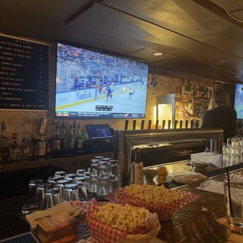 Jackpot - The image depicts the interior of a bar with a large flat-screen TV showing a hockey game. The bar counter has baskets of popcorn, glasses, and a menu. A bartender is seen serving drinks, and shelves with bottles are visible behind the counter. The setting appears casual and dimly lit, with a focus on sports viewing. - Dive Bars Near Me - Washington, DC, District of Columbia