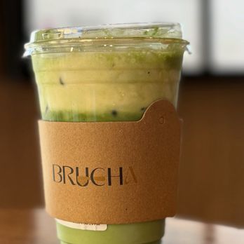 Brucha - Salted banana matcha - A green matcha drink in a clear plastic cup with a brown paper sleeve from Brucha, sitting on a wooden surface. The drink appears to have a layered or blended texture, with some darker specks visible. The background is blurred, suggesting an indoor setting with natural light. - Gelato Near Me - Tucson, Arizona
