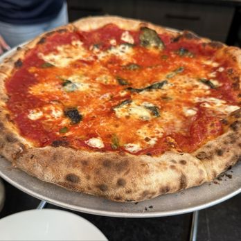 Britesmith Brewing - Buffalo - Cheese pizza - A cheese pizza with a golden-brown, slightly charred crust is displayed on a metal tray. The pizza has a layer of tomato sauce, melted cheese, and scattered basil leaves. The crust appears thick and airy with some darker spots indicating it was baked at high heat. The background shows part of a person and a dark interior setting. - New American Near Me - Buffalo, New York