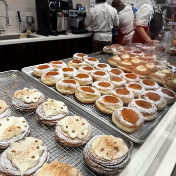 Kora - A variety of Filipino-inspired baked goods, including cream-filled pastries with bear-shaped decorations and caramel-topped doughnuts dusted with powdered sugar, are displayed on cooling racks behind a glass counter. The background shows a busy kitchen with staff preparing orders, and a coffee machine is visible on the counter. The setting appears to be a bakery or café specializing in Filipino-style pastries. - Filipino Near Me - Sunnyside, New York