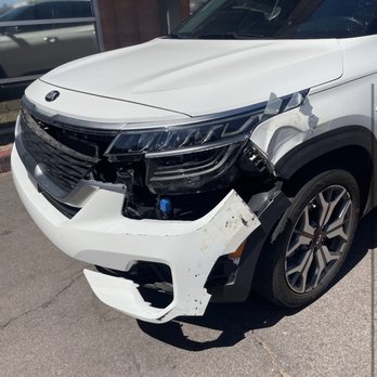 Master Tech Collision - A white Kia SUV with front-end damage is parked outside a building. The front bumper and headlight are missing or broken, with visible internal components and tape holding parts together. The vehicle is on a paved surface, and the background includes a glass window and a brick wall, indicating an outdoor setting near a business or repair shop. - Body Shops Near Me - Tempe, Arizona