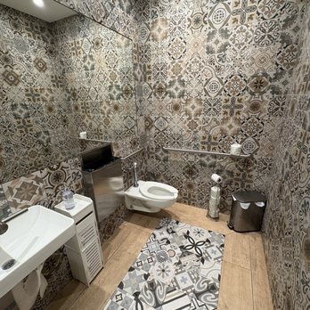 Rosetta Bakery - Nice restroom - A bathroom features patterned tile walls, a toilet, a small sink, a mirror, and a trash bin, with a wooden floor and a drain in the center. - Bakeries Near Me - Miami, Florida