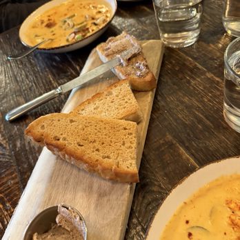 Forbidden Root - Beer bread - A wooden serving board holds slices of toasted bread and a small dish of spread. The table features two bowls of soup with a creamy appearance and garnished with herbs, along with glasses of water. The setting appears to be a casual dining environment with a rustic wooden table. - Brewpubs Near Me - Chicago, Illinois
