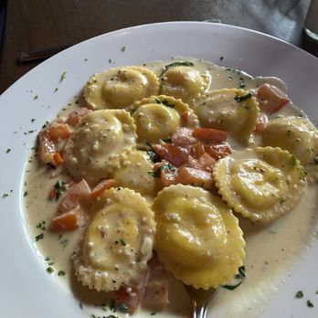 Giacomo's - A white plate holds ravioli pasta covered in a creamy white sauce, garnished with chopped herbs. Diced vegetables, including carrots and tomatoes, are visible around the pasta. The dish appears to be served in a restaurant setting with a dark wooden table surface. - Italian Near Me - Melrose, Massachusetts