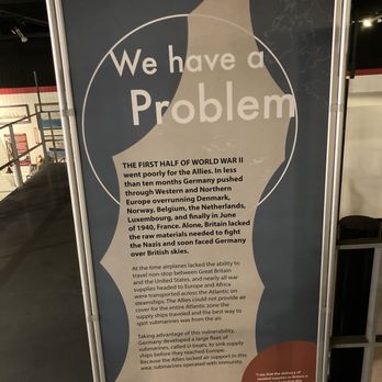 Marine Museum of Fall River - A tall, vertical sign displays historical information related to World War II, including details about Germany's military actions and submarine strategies. The sign features a silhouette of a submarine at the bottom and a quote in a red circle. It is situated indoors, likely within the Marine Museum of Fall River, with a background showing part of the museum interior and other exhibits. - Museums Near Me - Fall River, Massachusetts