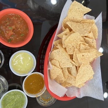 Los Braseros - A red basket lined with paper holds a serving of tortilla chips on a black table. Surrounding the basket are three small bowls containing different salsas and sauces, including red salsa, green salsa, and a yellowish sauce. The setting suggests a Mexican restaurant or casual dining environment with a focus on traditional snacks and condiments. - Mexican Near Me - Austin, Texas