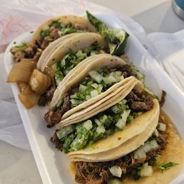 Carne Asada Tacos - Three tacos filled with seasoned beef, topped with chopped onions and cilantro, served on a foam tray. Accompanied by a pickled jalapeño and a roasted green pepper, with some pickled vegetables visible. The setting appears casual, likely from a food truck or casual eatery.