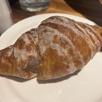 Cheddar's Scratch Kitchen - Sweet buttery croissant - A croissant with a shiny glaze and dusting of powdered sugar is placed on a white plate. The croissant appears freshly baked with a golden-brown exterior. The background includes a glass of water and a wooden table surface, indicating a dining environment. - American Near Me - York, Pennsylvania