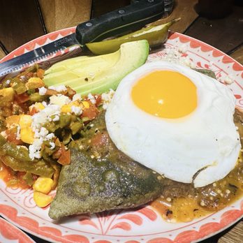 Mariscos San Pedro - Blue Corn Tlacoyo - A Mexican-style meal on a decorative ceramic plate includes a fried egg with a runny yolk, a stuffed tortilla covered in sauce, and a side of vegetable salsa with corn, peppers, and cheese. The plate is placed on a wooden table, with a knife and pickled jalapeños visible nearby. - Mexican Near Me - Chicago, Illinois