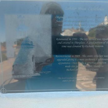 Lobster Point Lighthouse - Plaque - A glass plaque mounted on a wall contains text detailing the history and renovation of Lobster Point Lighthouse, including its original construction, renovation in 1993, and reconstruction in 2009. The reflection of a person holding a camera and some buildings and sky are visible in the glass, making the image a combination of the plaque's information and the photographer's reflection. - Landmarks & Historical Buildings Near Me - Ogunquit, Maine