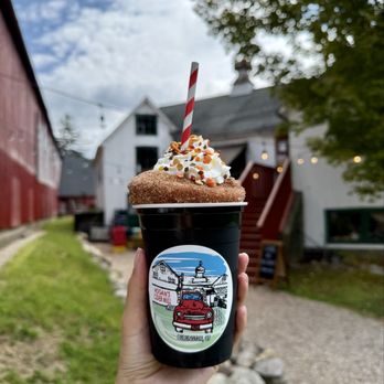 Hogan's Cider Mill - delightful donut ($11) - A person holds a cup topped with whipped cream, colorful sprinkles, and a striped straw, with a sticker of Hogan's Cider Mill in Burlington, CT. The background shows a historic white building with a steep roof, trees, and a partly cloudy sky, indicating an outdoor setting at the cider mill. - Venues & Event Spaces Near Me - Burlington, Connecticut