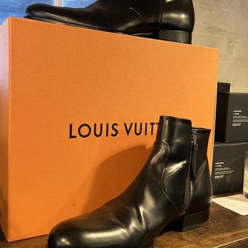 Stitch Switch - Mens resale - local down town Geneva - Two black leather ankle boots with a side zipper are positioned on a wooden surface, one in front and one on top of an orange Louis Vuitton box. The boots have a smooth, shiny finish and a low heel. The background includes a window and some retail packaging, indicating a store display for designer footwear. - Men's Clothing Near Me - Geneva, Illinois