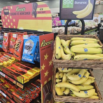QuickChek - The image shows a grocery store produce section with baskets of bananas on the right, labeled with a sign indicating they cost 99 cents each. To the left, there are shelves stocked with Reese's snack products, including Reese's Peanut Butter Cups and Hershey's popcorn, with promotional signage. The store has a ceiling with bright, square lighting panels and other products visible in the background. - Gas Stations Near Me - Hamilton Township, New Jersey
