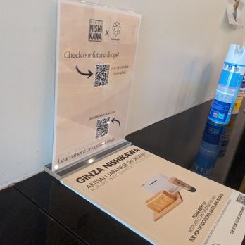 Sunright Tea Studio - The image shows a promotional display on a table for Ginza Nishikawa, an artisan Japanese rice cracker brand. The display includes a printed sign with the brand name, product image, and QR codes for future product drops and contact information. The sign is placed on a black surface, with a clear acrylic stand holding an additional informational sheet. To the right, there are bottles of cleaning or disinfectant spray, and a wall with some posters or notices. - Bubble Tea Near Me - Fullerton, California