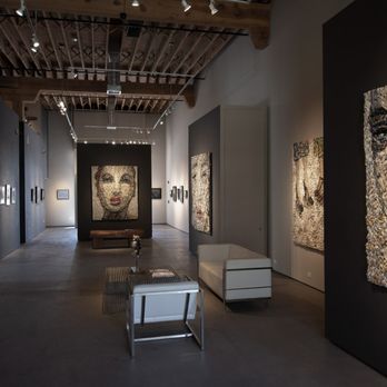 EVOKE Contemporary - Gugger Petter woven newspaper artwork at Evoke Contemporary Gallery - The image shows an art gallery interior with high ceilings and exposed wooden beams. The gallery features textured portrait artworks mounted on dark and light-colored walls, with a focus on detailed, mixed-media faces. There are seating arrangements, including a white sofa and a small table, in the foreground. The space is well-lit with track lighting highlighting the artworks, and the overall atmosphere is modern and minimalistic. - Art Galleries Near Me - Santa Fe, New Mexico