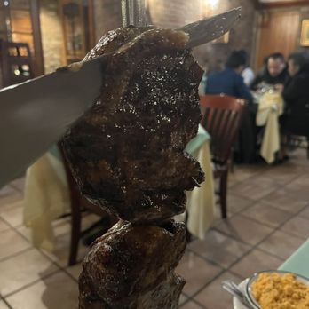 Fernandes steakhouse - Cap Steak (picanha) - A close-up of a skewer with grilled meat, held by a hand inside a restaurant with diners in the background. - Portuguese Near Me - Newark, New Jersey