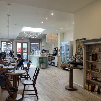 Loaf on Paper - The image shows the interior of a Brooklyn cafe with a high ceiling and wooden flooring. Customers are seated at round tables, working on laptops. The cafe has a curved counter with a barista, a beverage fridge, and decorative lighting fixtures, including a paper lantern and string lights. The space is well-lit with natural light coming through the front windows and doors, creating a welcoming atmosphere. - Cards & Stationery Near Me - Brooklyn, New York