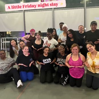 DivaDance - Friday Night Dance Class - A group of women, dressed in casual and dance attire, are gathered in a dance studio, smiling and posing for a photo. They are kneeling or sitting on the floor, with some standing behind. The background features large windows and a poster of a woman with blue hair and a caption that reads 'A little Friday night slay.' The setting suggests a dance class or social event at DivaDance in Brooklyn, NY. - Dance Studios Near Me - Brooklyn, New York