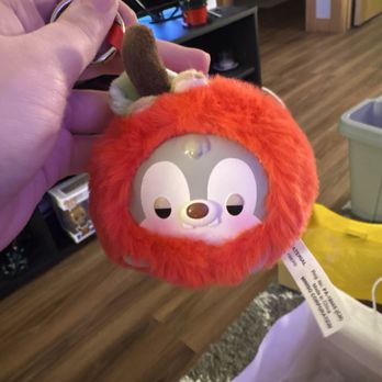 Miniso - Fruit Disney blind box - A plush Disney character toy with a face inside a red furry cover, attached to a keychain, is held by a person's hand. The toy has a character face with large eyes and a small nose, and the furry cover resembles a pumpkin or fruit. The background shows a living room with a TV, a shelf, and some household items. - Toy Stores Near Me - Bloomington, Minnesota
