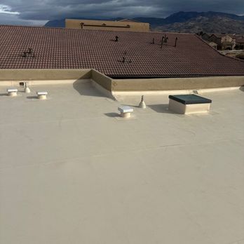 AJ's Professional Contracting - 3-Shiny new roof - The image shows a flat rooftop with several vents and a small access hatch, all secured with white coverings. The roof surface appears smooth and well-maintained. In the background, there are neighboring rooftops with red tiles, and beyond that, a mountain range under a cloudy sky. The scene suggests a residential or commercial building in a mountainous area. - Roofing Near Me