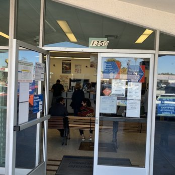 US Post Office - Worst post office ever! Very slow and very inefficient - The image shows the entrance of a post office with glass sliding doors. Inside, several customers are seated and standing near the service counter. The interior ceiling has fluorescent lighting, and various signs are posted on the glass doors and windows. The building number 1350 is displayed above the entrance. Outside, parking and other storefronts are partially visible. - Post Offices Near Me - Fullerton, California