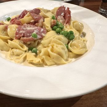 La Barra - Riverside - A white plate holds tortellini pasta with green peas, slices of ham, and a creamy sauce, set on a wooden table with a glass of water nearby. - Pizza Near Me - Riverside, Illinois
