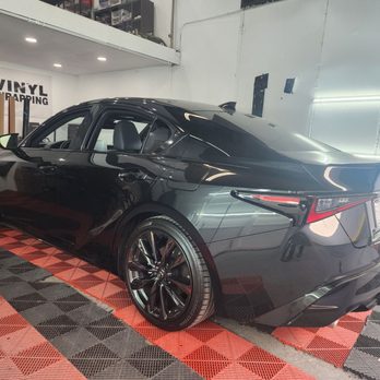 Wrap Clinic - Looks great!!! - A black sedan with a modern, aerodynamic design is parked inside a garage with red and black checkered flooring. The vehicle has tinted windows, black alloy wheels, and a rear spoiler. The garage features white walls, some wall-mounted equipment, and a partially visible upper storage area with various items. The lighting reflects off the car's surface, highlighting its glossy finish. - Vehicle Wraps Near Me - Fullerton, California