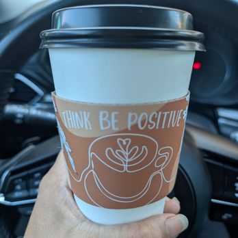 Inspired Brew - Reminder with your coffee - A person holds a coffee cup with a cardboard sleeve that has the message 'THINK BE POSITIVE' and a smiley face illustration. The cup has a black lid and is held in front of a car's steering wheel, indicating the photo was taken inside a vehicle. The background shows part of the dashboard and windshield, with natural light coming through. - Coffee & Tea Near Me - Dallas, Texas