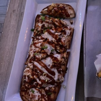 The Local - Hawaiian pizza app - A rectangular white plate holds several slices of pizza topped with sauce, cheese, chopped green onions, and other ingredients. The pizza slices are arranged in a row on a wooden table, with one piece slightly separated from the rest. The setting appears to be a casual dining environment, possibly outdoors or in a rustic restaurant. - Cocktail Bars Near Me - Babylon, New York