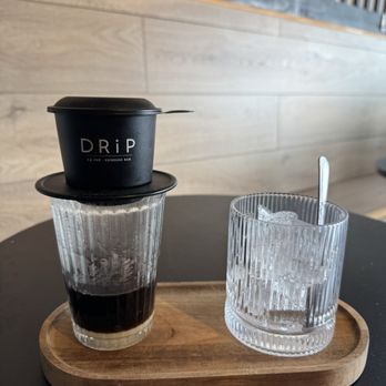 DRiP Cà Phê - A black coffee cup with a lid is stacked on a glass of dark coffee, placed on a wooden tray alongside a glass of ice water with a spoon. - Coffee & Tea Near Me - Annandale, Virginia