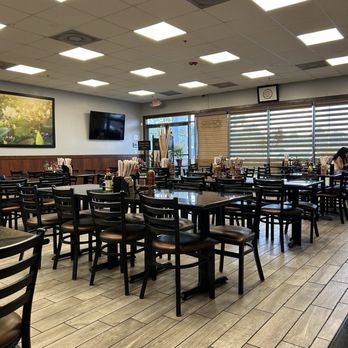 Pho 79 - The image shows an interior of a Vietnamese restaurant with multiple black tables and matching chairs arranged in rows. The space has a wood-paneled wall along one side, with a large framed picture and a wall-mounted TV. Large windows with horizontal blinds allow natural light into the room. The ceiling has a grid of fluorescent lights, and the floor is covered with wood-look tiles. Condiments and utensils are placed on the tables, and a person is visible seated at one of the tables in the background. - Vietnamese Near Me - Miami, Florida