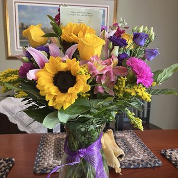 Armstrong Flowers - A vibrant bouquet of mixed flowers, including sunflowers, yellow roses, pink lilies, purple lisianthus, and greenery, is arranged in a clear glass vase tied with a purple ribbon. The bouquet is set on a wooden dining table with woven placemats and cloth napkins. In the background, a framed picture and a card with a Valentine's Day message are visible on the wall. - Florists Near Me - Fort Wayne, Indiana