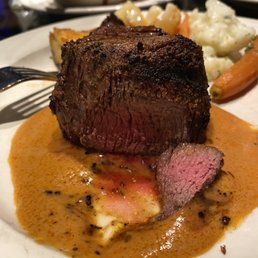 8 Oz. Tender Filet Mignon - A close-up of a plate with a cooked filet mignon topped with Diane sauce, showing a pink interior and a seared exterior. The dish is served with mashed potatoes, carrots, and a vegetable medley in the background. The presentation highlights the steak and its accompaniments, typical of a steakhouse meal.