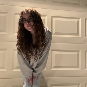 Roots Salon - A woman with long, curly hair and glasses is standing in front of a closed garage door, wearing a light-colored hoodie and pants. - Hair Salons Near Me - Albuquerque, New Mexico