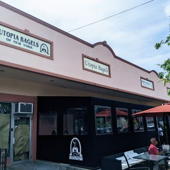 Utopia Bagels - Outside - The photo depicts the exterior of Utopia Bagels of New York, featuring a pink building with signage displaying the business name. There is an outdoor seating area with tables, chairs, and umbrellas, where several people are seated. The storefront includes a black section with a logo and a glass door with an 'Open' sign. The scene is set during daytime with a partly cloudy sky and some greenery visible on the right side. - Bakeries Near Me - Whitestone, New York