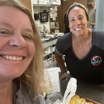Maxim's Pizza - Customer service! Very best! - The image shows two women smiling at the camera inside a pizza restaurant. The woman on the left has blonde hair and is wearing a gray top, while the woman on the right has dark hair, a headband, and is wearing a black shirt with a Maxims logo. There are pizza boxes, a drink, and a menu on the counter in front of them. The background includes kitchen equipment, shelves with boxes, and various restaurant supplies, indicating a casual dining environment. - Pizza Near Me - Mentor, Ohio