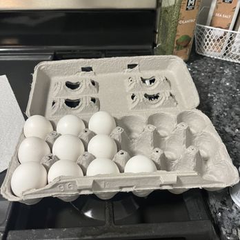 Safeway - A carton containing twelve white eggs is positioned on a stovetop in a kitchen setting. The carton is open, revealing the eggs arranged in two rows. In the background, there are spice jars labeled cilantro and sea salt, along with a white basket holding additional containers. The surface around the stovetop is a dark, speckled countertop. - Grocery Near Me - Concord, California